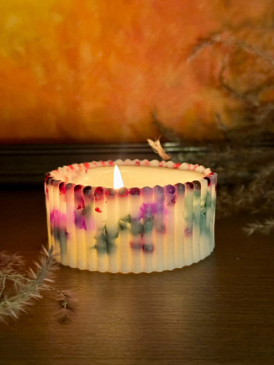 Degoša, balta rievota dizaina svece ar iestrādātām violetām un zaļām ziedlapiņām mājīgā apgaismojumā uz koka virsmas