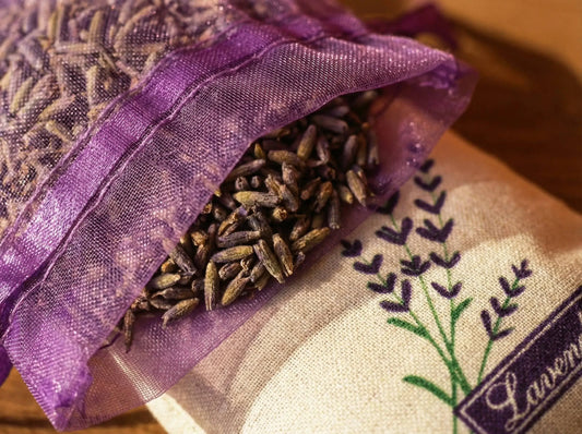 Kaltētu lavandas ziedu tuvplāns, kas birst ārā no violeta organzas maisiņa uz lina auduma maisiņa ar lavandas izšuvumu