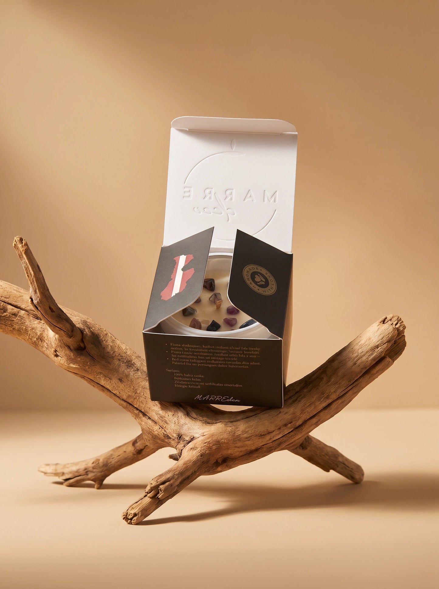 Daļēji atvērta MarreDeco sveces melnā kastīte ar Latvijas kontūru, atsedzot dabīgā vaska sveci ar violetiem un tumšiem kristāliem, novietota uz dekoratīva koka zara.
