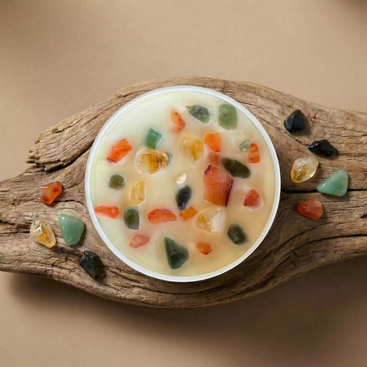 Balta metāla kārbiņa ar nodoma sveci "Dzīvespriekam", dekorēta ar krāsainiem kristāliem, novietota uz gaiša, poraina akmens pamatnes saulainā telpā ar palmu lapu ēnu rakstiem.