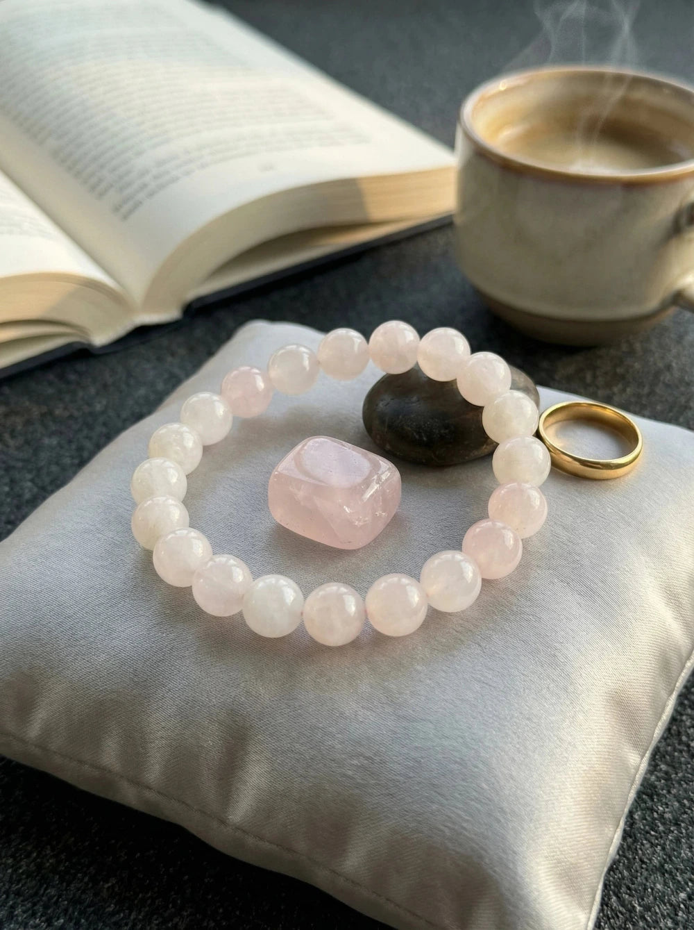 Gaiši rozā kristāla pērlīšu rokassprādze blakus zelta gredzenam un kūpošai kafijas tasei uz tumša fona