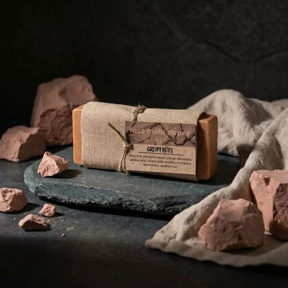 Rozā mālu ziepes ar kazas pienu "Greipfrūts" uz tumša akmens paliktņa, dekorētas ar dabīgiem rozā māla gabaliem un lina audumu.