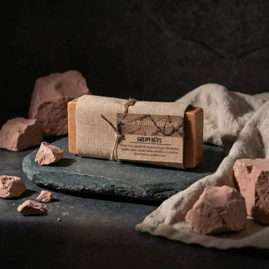 Rozā mālu ziepes ar kazas pienu "Greipfrūts" uz tumša akmens paliktņa, dekorētas ar dabīgiem rozā māla gabaliem un lina audumu.