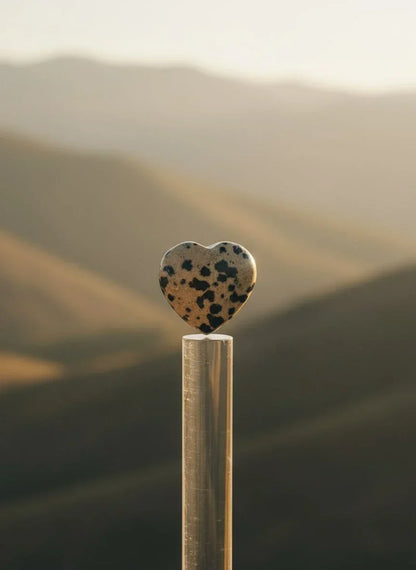 Dalmācijas jašmas sirds formas akmens, kas novietots uz caurspīdīga statīva uz miglainu kalnu un saulrieta fona