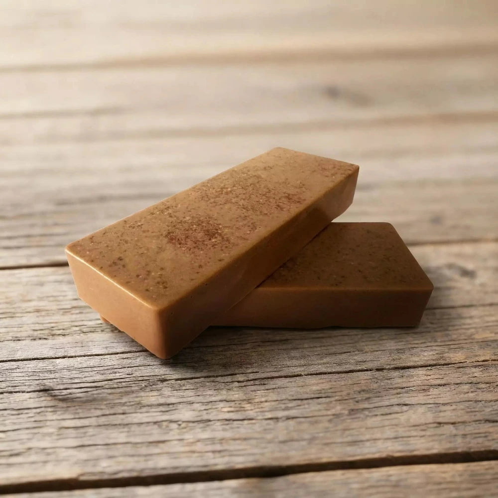 Divi taisnstūra formas brūni, dabīgas izcelsmes ziepju stienīši ar smalku tekstūru, novietoti krusteniski viens uz otra uz veca koka dēļu fona.