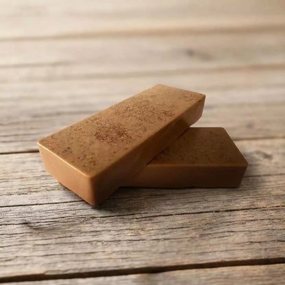 Divi taisnstūra formas brūni, dabīgas izcelsmes ziepju stienīši ar smalku tekstūru, novietoti krusteniski viens uz otra uz veca koka dēļu fona.