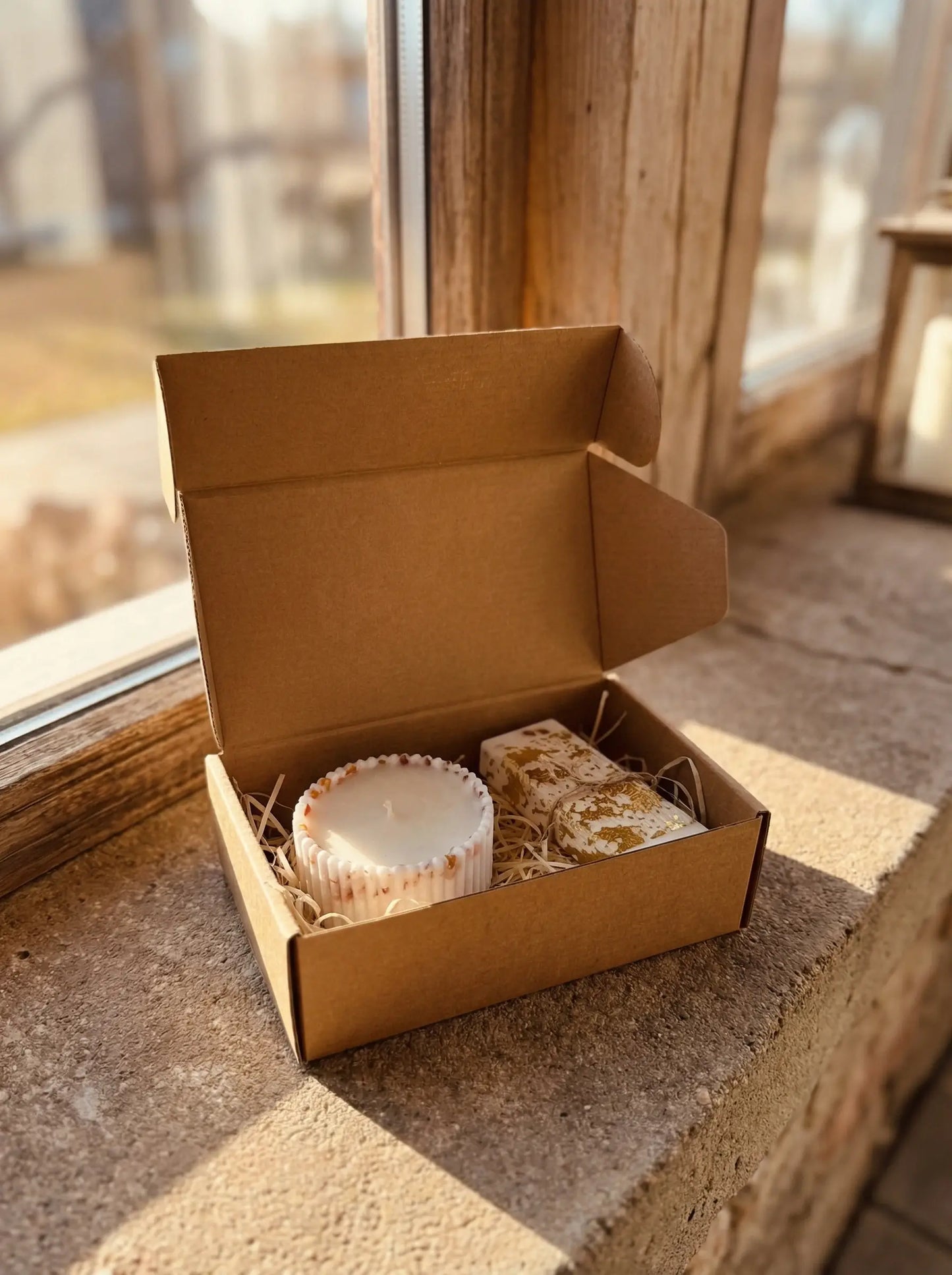 Atvērta brūna kartona dāvanu kaste ar dekoratīvu sveci un vaska plāksnīti uz saules apspīdētas palodzes – dabīga dāvana no MarreDeco.