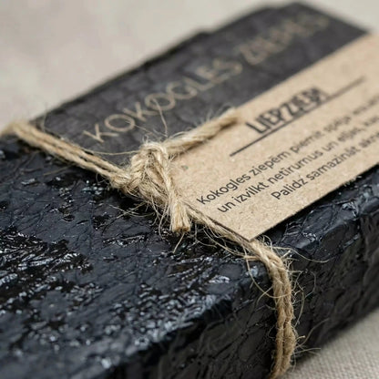 Melnas kokogles ziepes "Citronzāle", novietotas uz lina auduma blakus lielam kokogles gabalam un svaigiem citronzāles kātiem