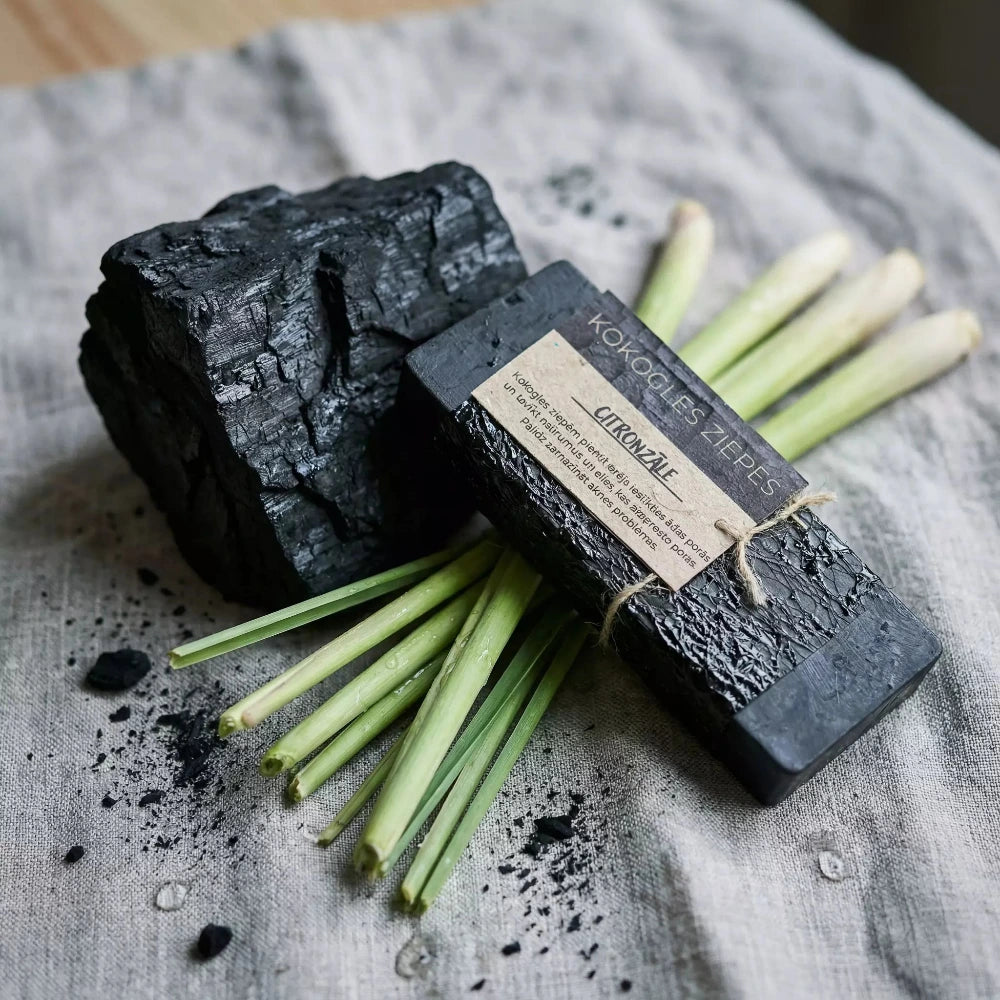 Melnas kokogles ziepes "Citronzāle", novietotas uz lina auduma blakus lielam kokogles gabalam un svaigiem citronzāles kātiem