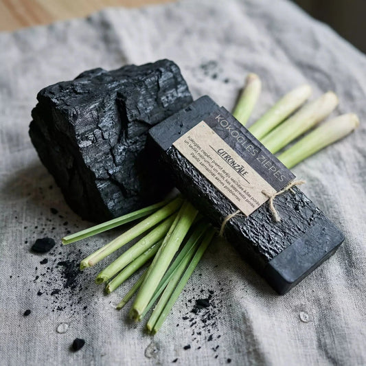 Melnas kokogles ziepes "Citronzāle", novietotas uz lina auduma blakus lielam kokogles gabalam un svaigiem citronzāles kātiem