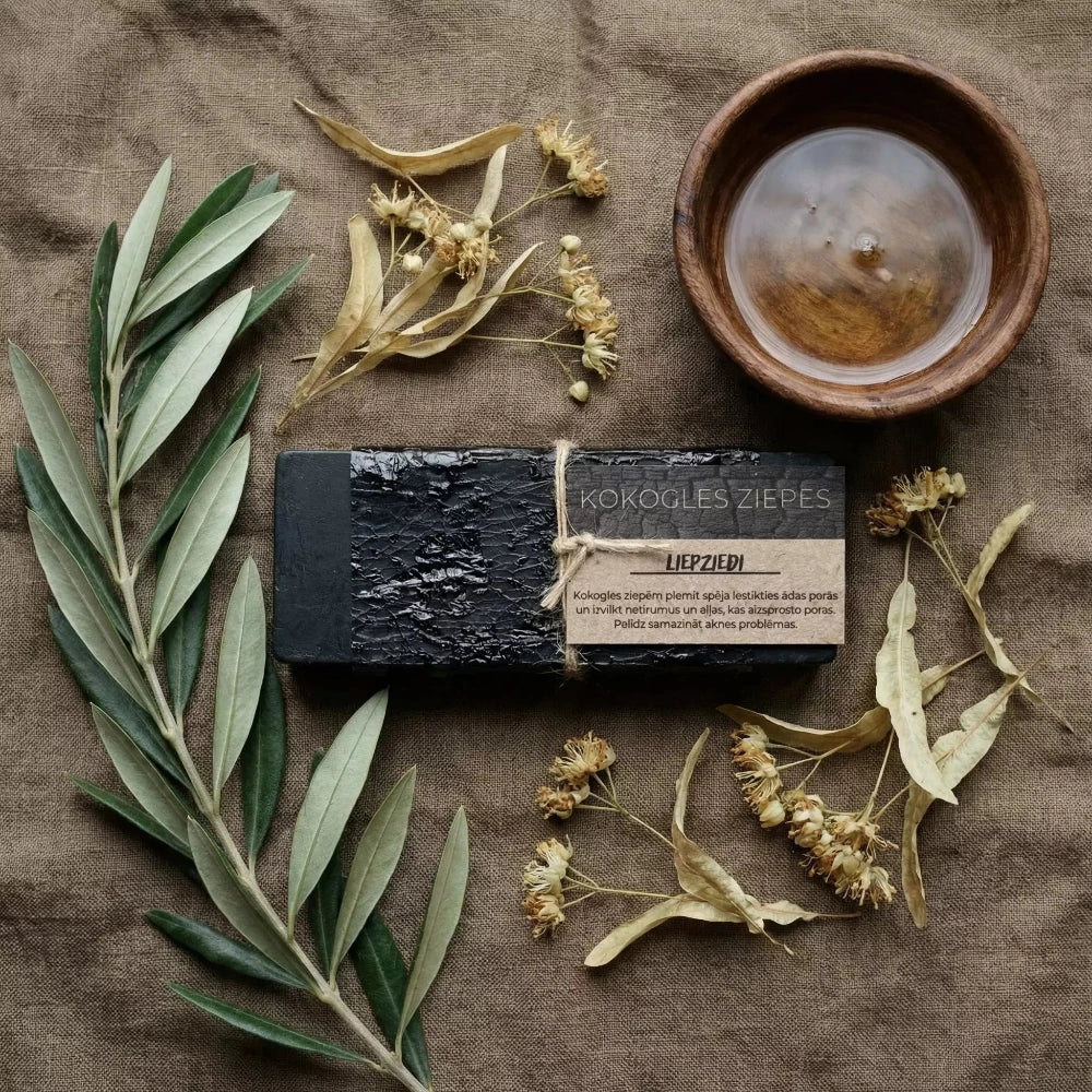 Melnas kokogles ziepes "Liepziedi" ar reljefu tekstūru, novietotas uz lina auduma blakus olīvzaram, kaltētiem liepziediem un koka bļodiņai ar eļļu.