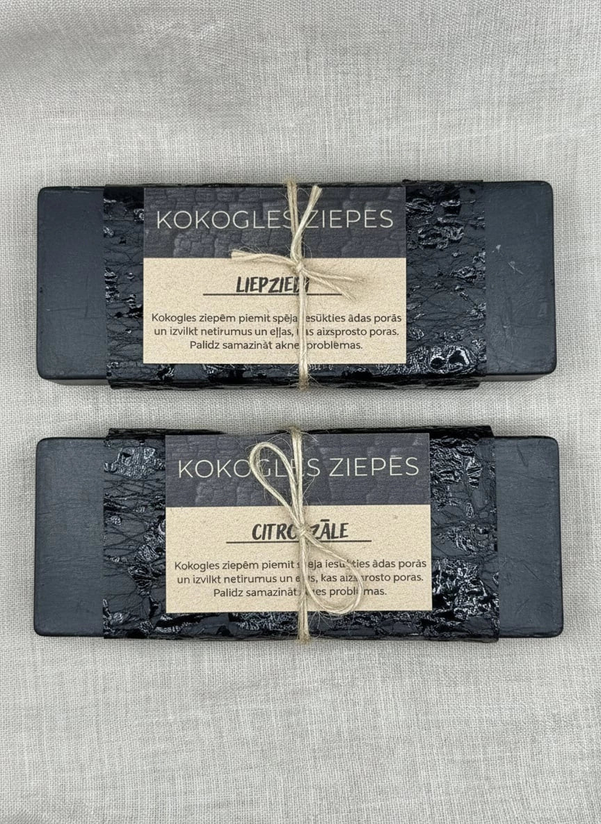 Divi melni kokogles ziepju gabali — "Liepziedi" un "Citronzāle" — ar reljefa tekstūru, ietīti dekoratīvā papīrā un pārsieti ar dabīgu lina auklu uz pelēka auduma fona