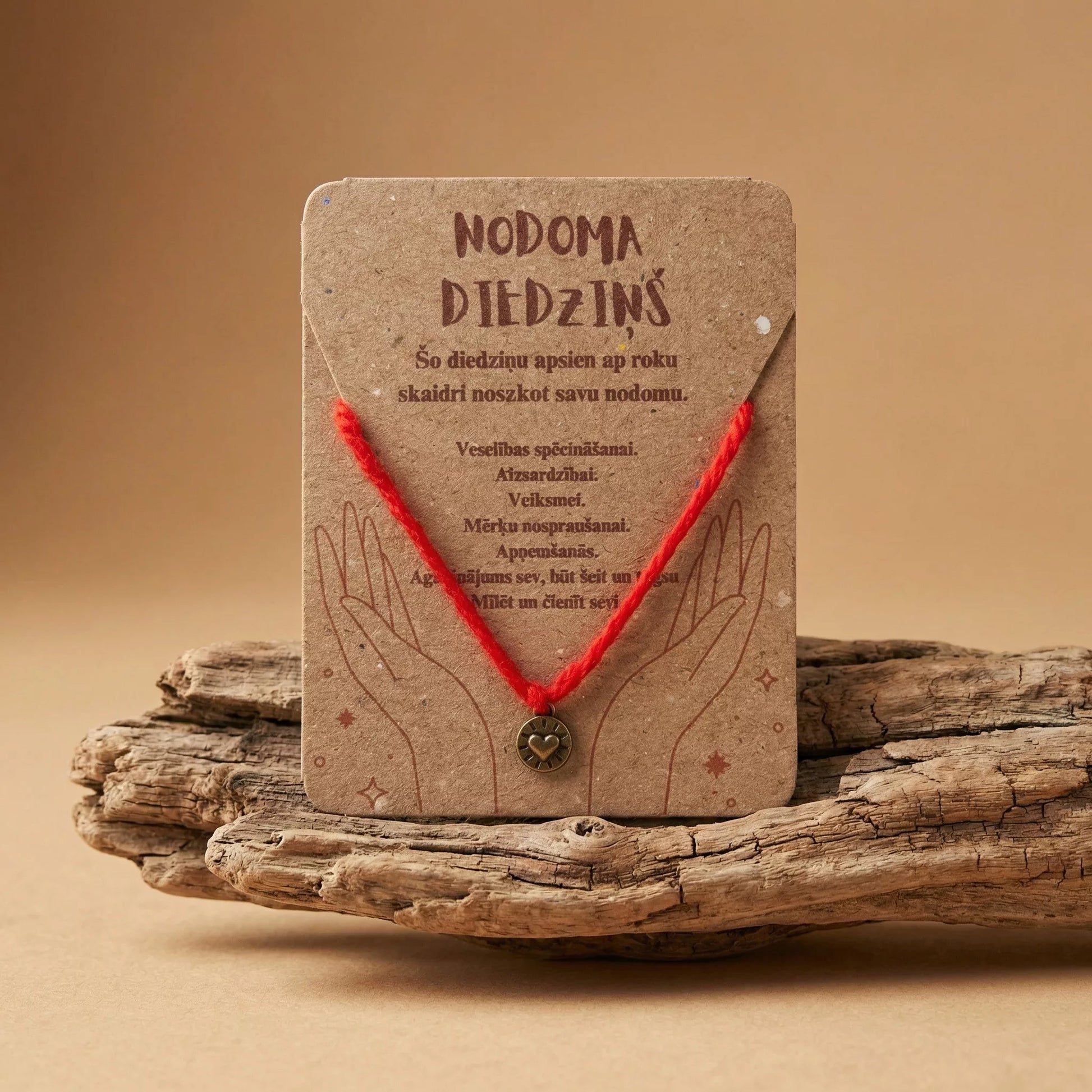 Sarkans nodoma diedziņš ar bronzas krāsas sirds piekariņu uz kartītes "Nodoma diedziņš" uz gaiša fona ar koku, marredeco