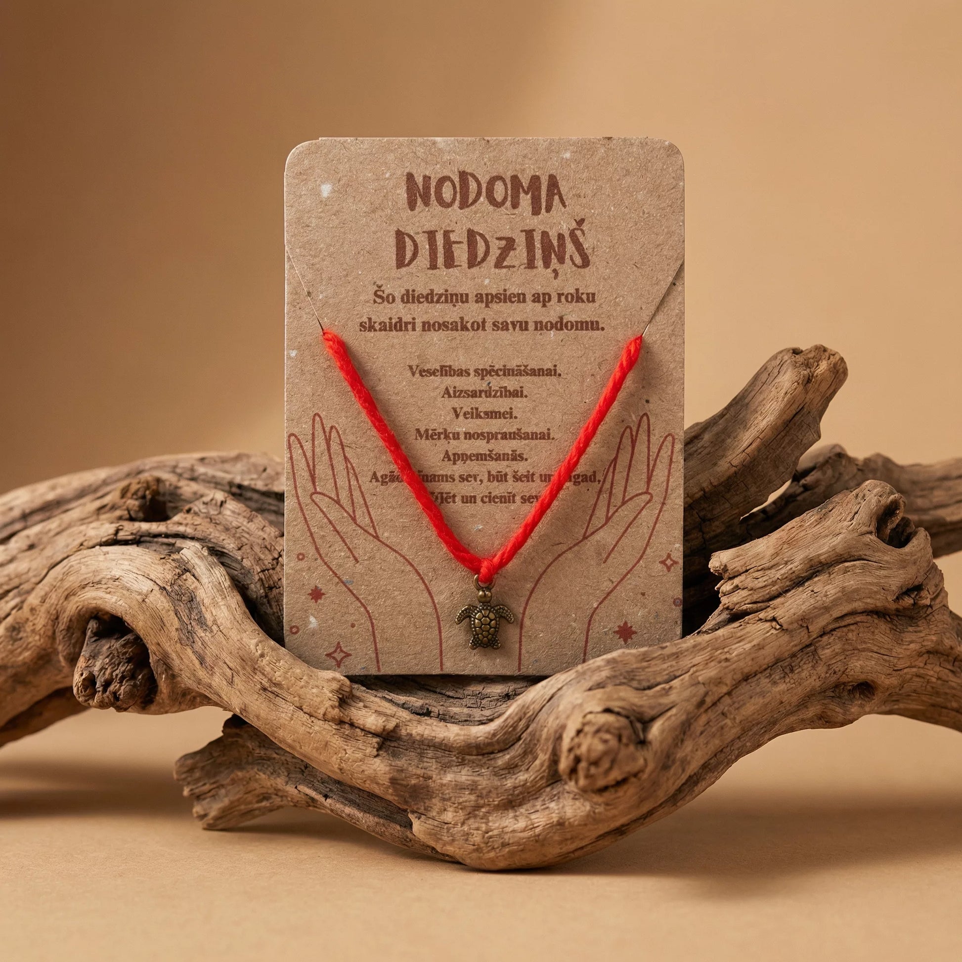 Sarkans nodoma diedziņš ar bronzas krāsas bruņurupuča piekariņu uz kartītes, novietots uz dekoratīva koka zara, marredeco