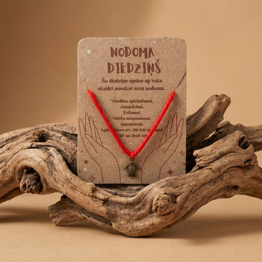 Sarkans nodoma diedziņš ar bronzas krāsas bruņurupuča piekariņu uz kartītes, novietots uz dekoratīva koka zara, marredeco