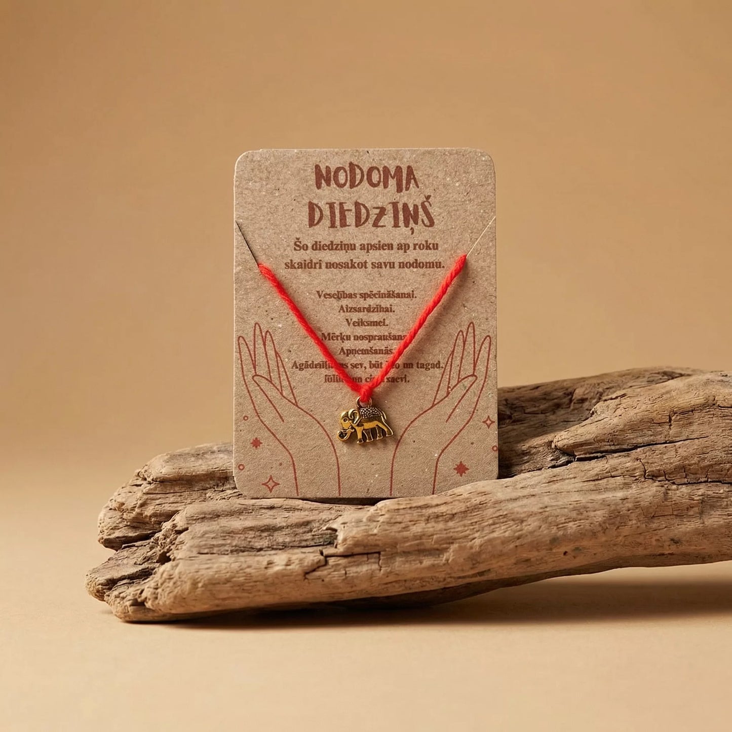 Sarkans nodoma diedziņš ar zeltītu ziloņa piekariņu uz kartītes "Nodoma diedziņš", novietots uz dabīga koka zara, marredeo