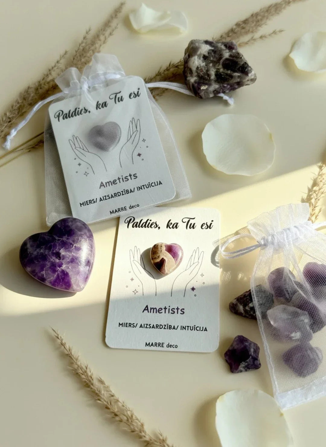 Ametista sirds formas akmens uz kartītes ar uzrakstu "Paldies, ka Tu esi" un nozīmi "Miers / Aizsardzība / Intuīcija". Fonā redzams balts organzas maisiņš, neapstrādāti ametista kristāli un dekoratīvas ziedlapiņas