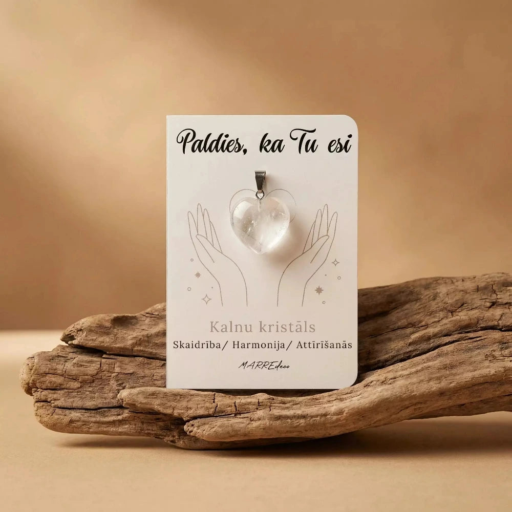 Caurspīdīgs kalnu kristāla sirds formas kulons uz baltas kartītes ar uzrakstu "Paldies, ka Tu esi" un nozīmi "Skaidrība / Harmonija / Attīrīšanās". Kartīte novietota uz dekoratīva koka zara