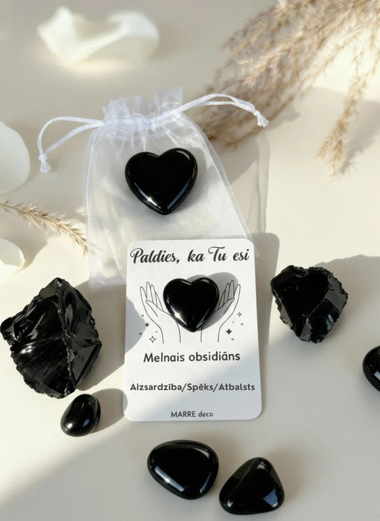 Melnā obsidiāna sirds formas akmens uz kartītes ar uzrakstu "Paldies, ka Tu esi". Apkārt izvietoti neapstrādāti obsidiāna kristāli, balti ziedi un balts organzas maisiņš gaišā, estētiskā kompozīcijā.