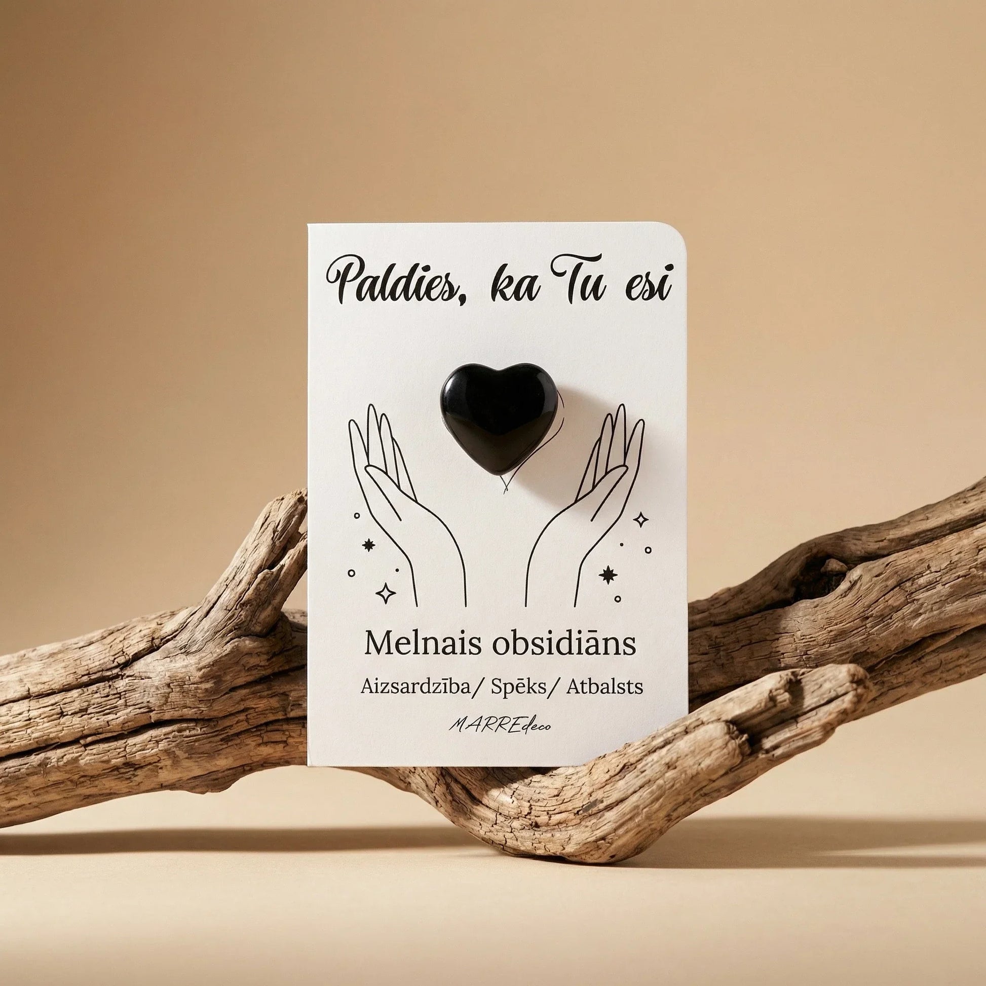 Melnā obsidiāna sirds formas akmens uz baltas kartītes ar uzrakstu "Paldies, ka Tu esi" un nozīmi "Aizsardzība / Spēks / Atbalsts". Kartīte novietota uz dabiska koka zara uz silta, bēša fona