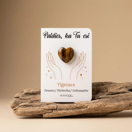 Brūna tīģeracs sirds formas akmens uz baltas kartītes ar uzrakstu "Paldies, ka Tu esi" un nozīmi "Drosme / Pārliecība / Gribasspēks". Kartīte novietota uz dabiska koka zara uz silta bēša fona