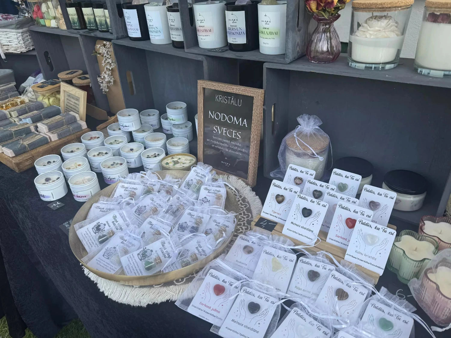 marredeco tirdziņa stends ar plašu roku darba produktu klāstu: kristālu nodoma svecēm metāla kārbiņās, aromātiskajiem vaskiem sirsniņu formā un dabīgo kosmētiku uz tumša auduma fona