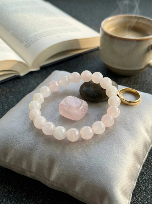 Gaiši rozā kristāla pērlīšu rokassprādze blakus zelta gredzenam un kūpošai kafijas tasei uz tumša fona