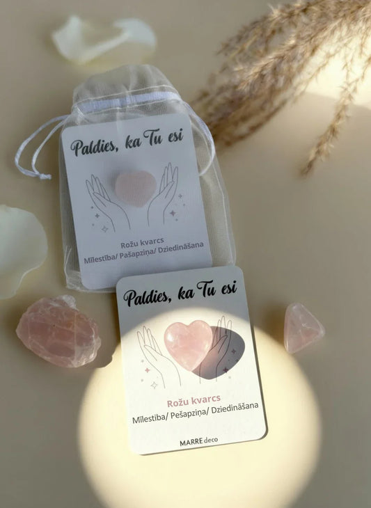 Rozā kvarca sirsniņa piestiprināta pie baltas kartītes ar uzrakstu "Paldies, ka Tu esi", ko papildina apraksts par mīlestību un dziedināšanu. Blakus redzams balts organzas maisiņš un neapstrādāti kristāli siltā apgaismojumā, marredeco