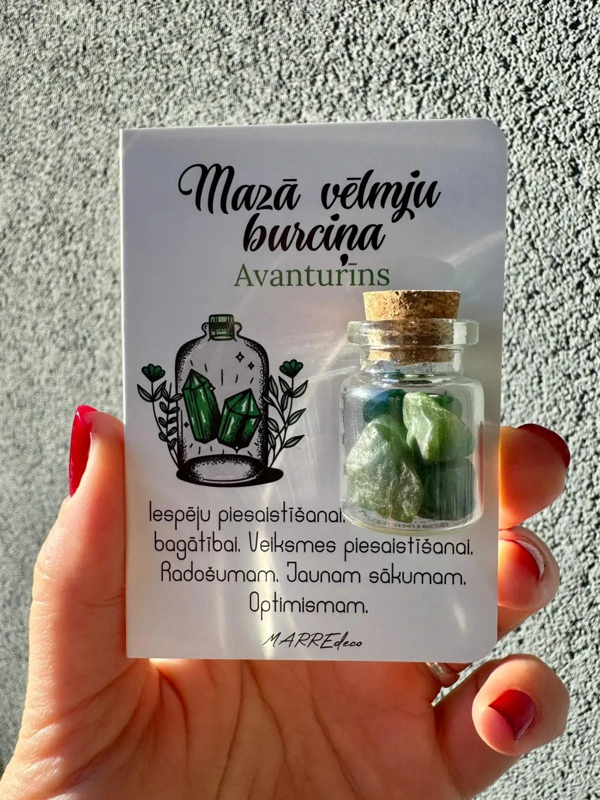 MarreDeco "Mazā vēlmju burciņa" ar zaļā avanturīna kristāliem iespēju piesaistīšanai, radošumam un optimismam, turēta rokā pret pelēku fonu