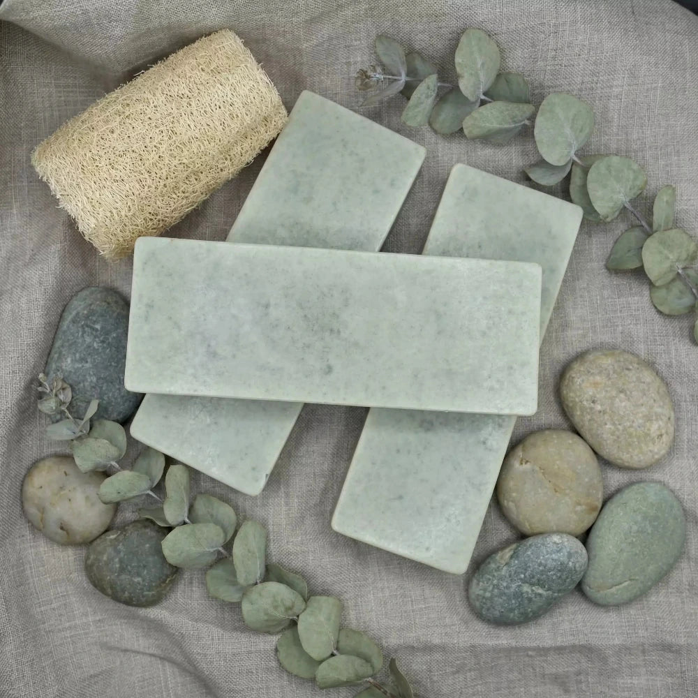 Trīs gaiši zaļas māla ziepes, sakārtotas uz lina auduma fonā ar eikalipta zariņiem, dabiskiem oļiem un lufas sūkli.