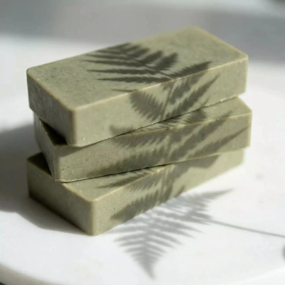 Trīs zaļā māla ziepju gabaliņi sakrauti kaudzītē, uz kuriem krīt estētiska papardes lapas ēna saules gaismā.