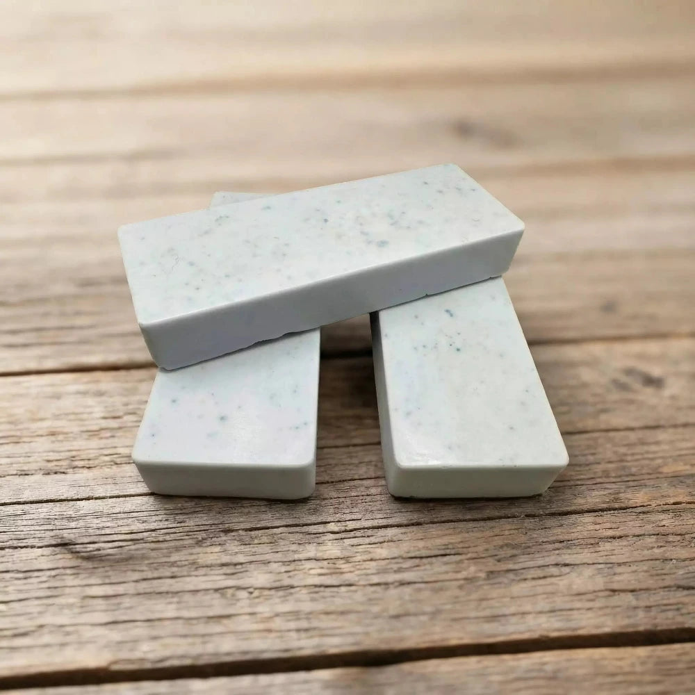 Trīs gaiši zilas māla ziepju plāksnītes ar tumšākiem punktiem, sakrautas kaudzītē uz koka virsmas.